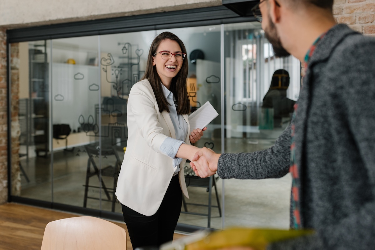 Smiling professional woman shakes hands with a colleague in a modern office while holding a notebook.